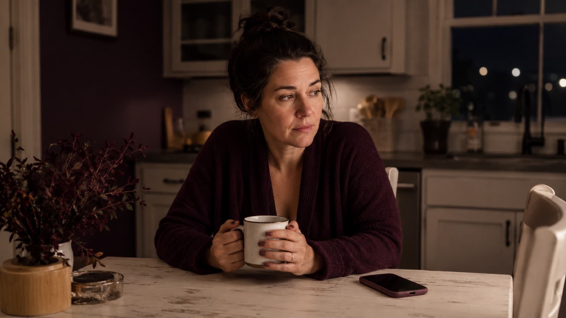 A woman sits at a kitchen table late at night, holding a mug and looking out the dark window, a phone resting nearby.