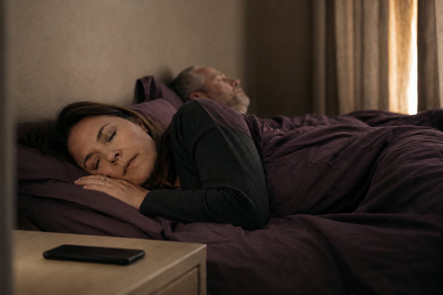 Two people sleep soundly at night while a phone rests dark and silent on the nightstand beside them.