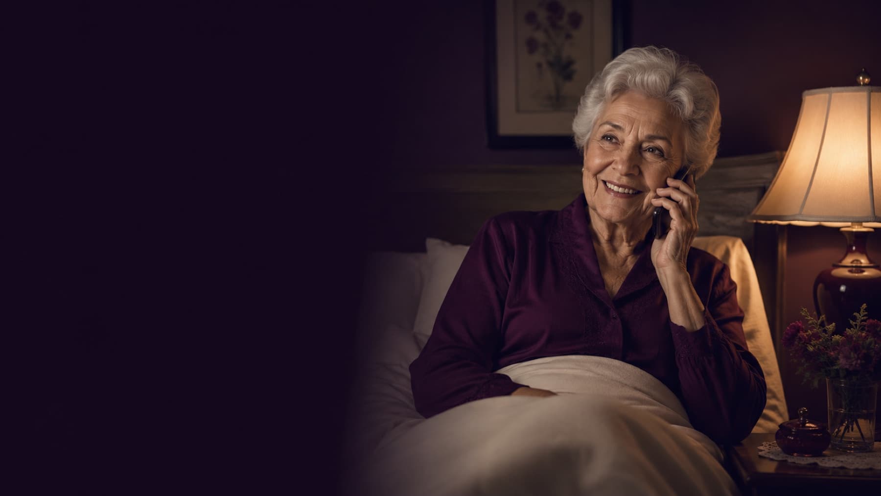 An older woman sits up in bed at night, smiling on the phone beside a warm bedside lamp.