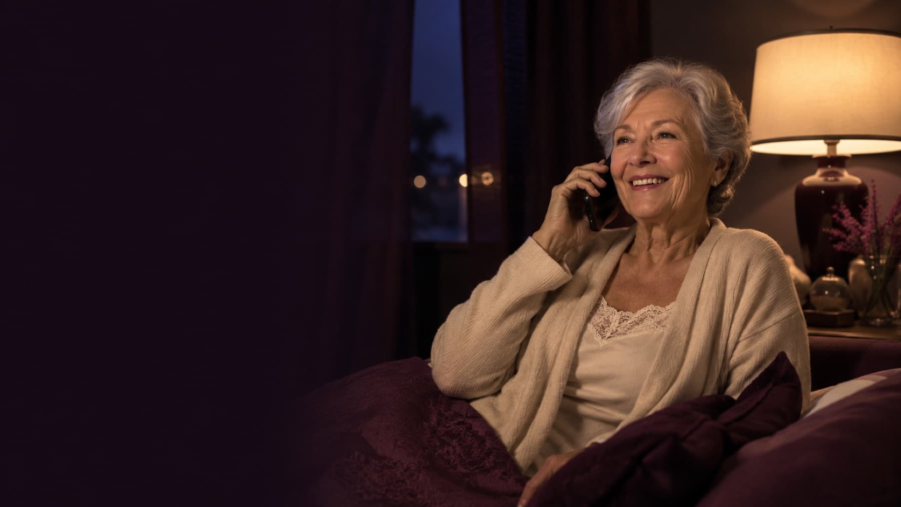 An older woman rests against pillows at night, smiling on the phone, with a curtained window and lamp glowing behind.